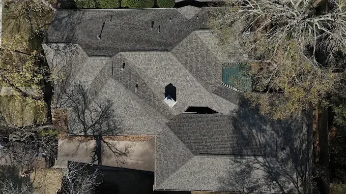 Overhead view of a multi-faceted house with a complex, shingled roof and surrounding bare trees.