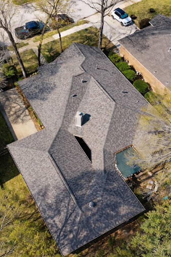 Overhead view of a house with a complex shingled roof, trees, and parked cars.