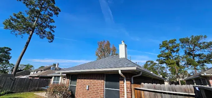 A blue sky with a contrail over a brick house and tall trees.