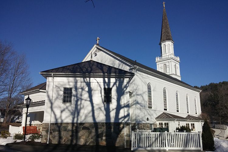 A white church with a steeple is surrounded by snow and trees.