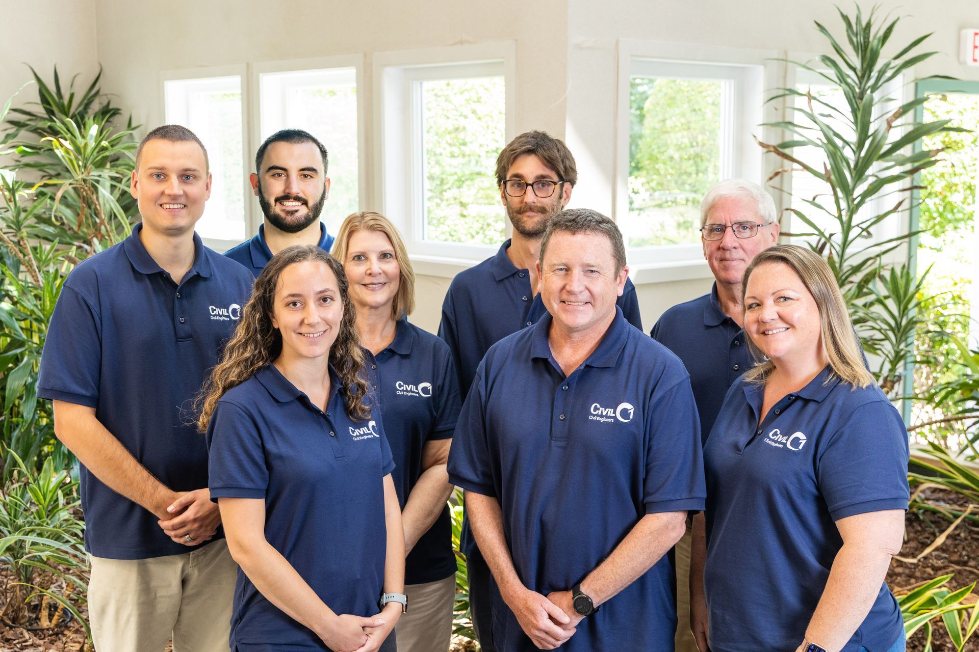 Group of people in matching blue shirts pose in front of a window, smiling.