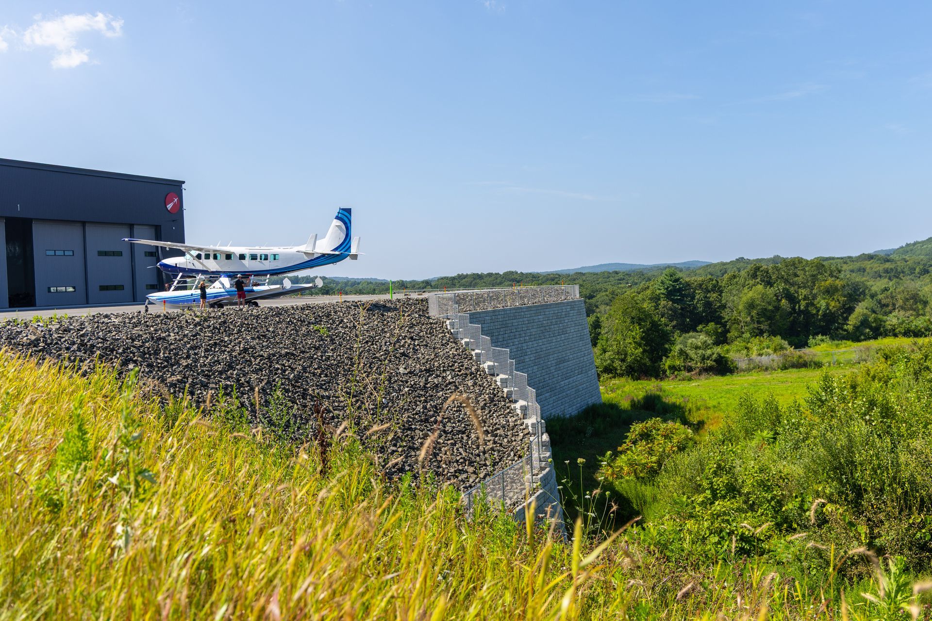 Airplane parked outside a hangar on a hillside, overlooking trees and a bright blue sky.