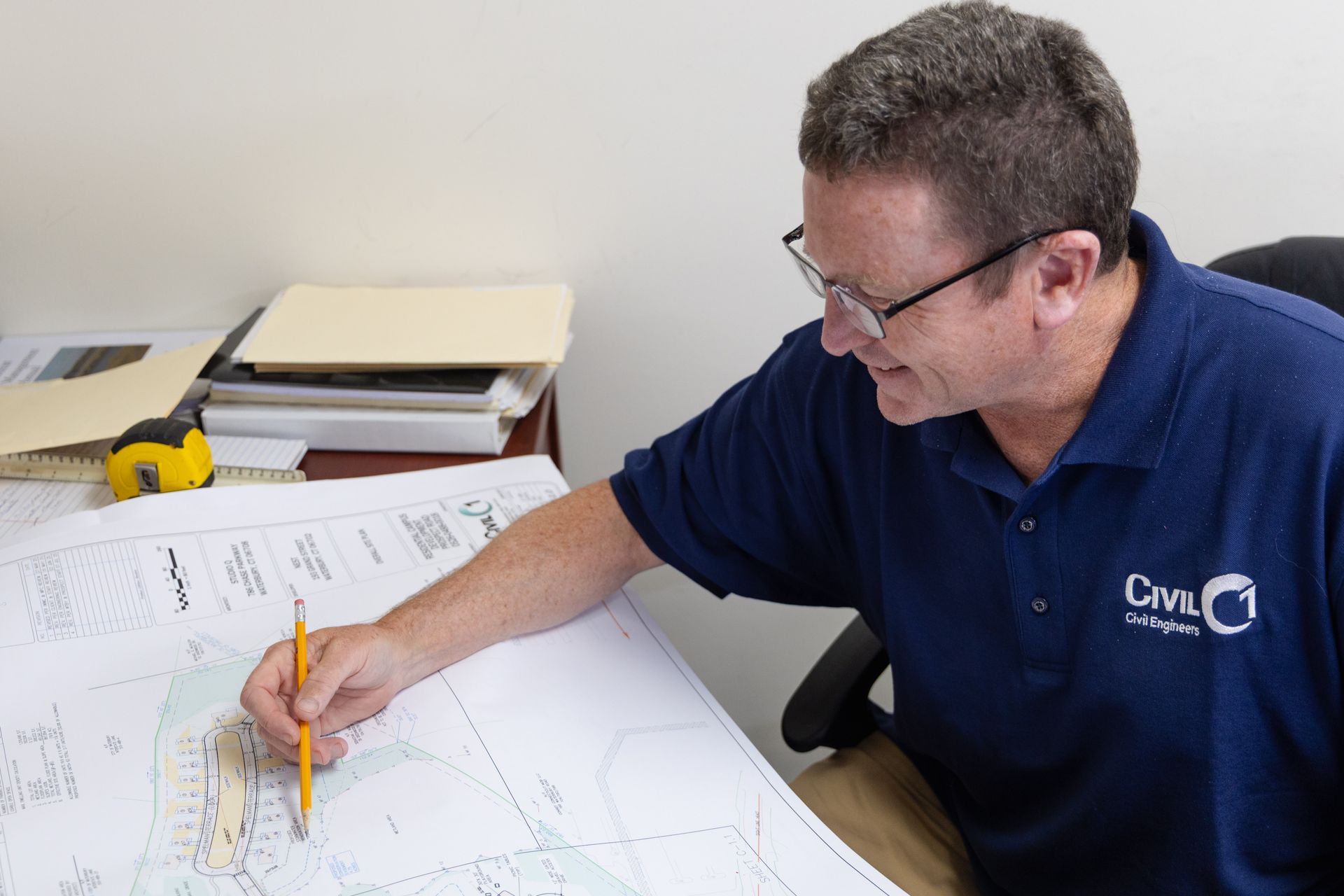 Man in a blue shirt with glasses, looking at blueprints with a pencil at a desk.