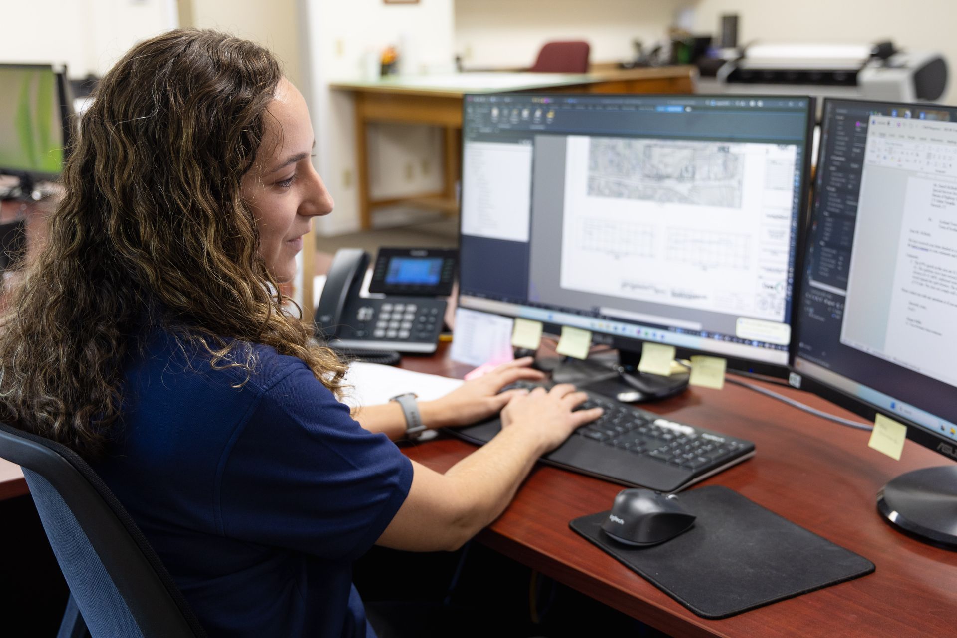 Woman working at a desk with three computer monitors, typing on a keyboard, and looking at the screens.