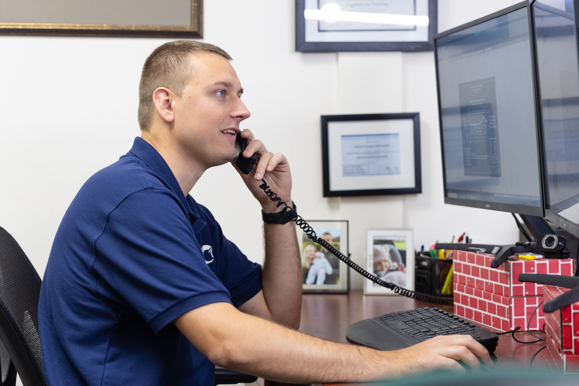 Man in blue shirt, on the phone at desk, looking at a computer screen. Office setting.