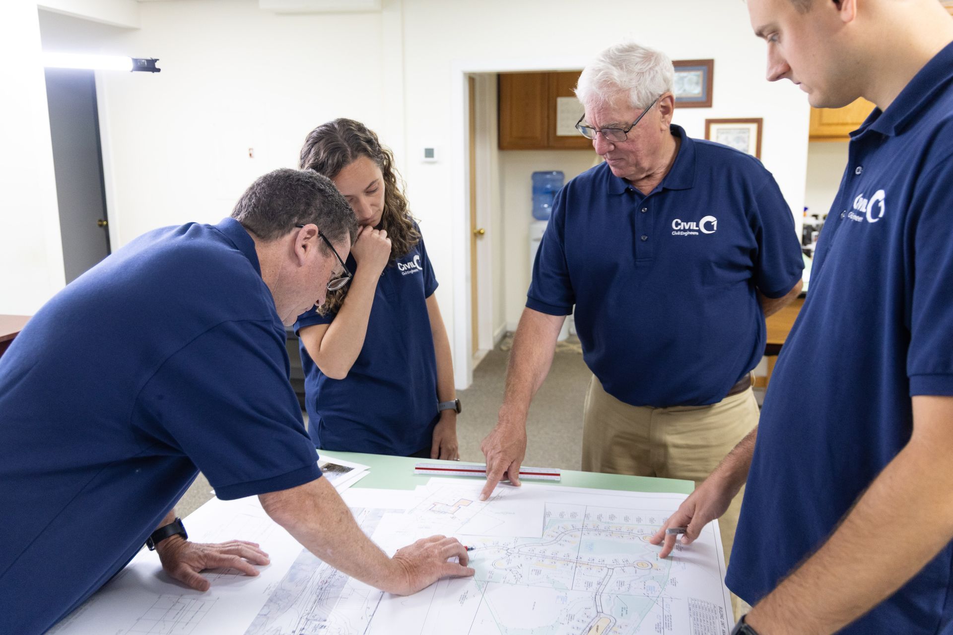 Four people in blue shirts examine a blueprint spread on a table in an office, pointing and discussing.