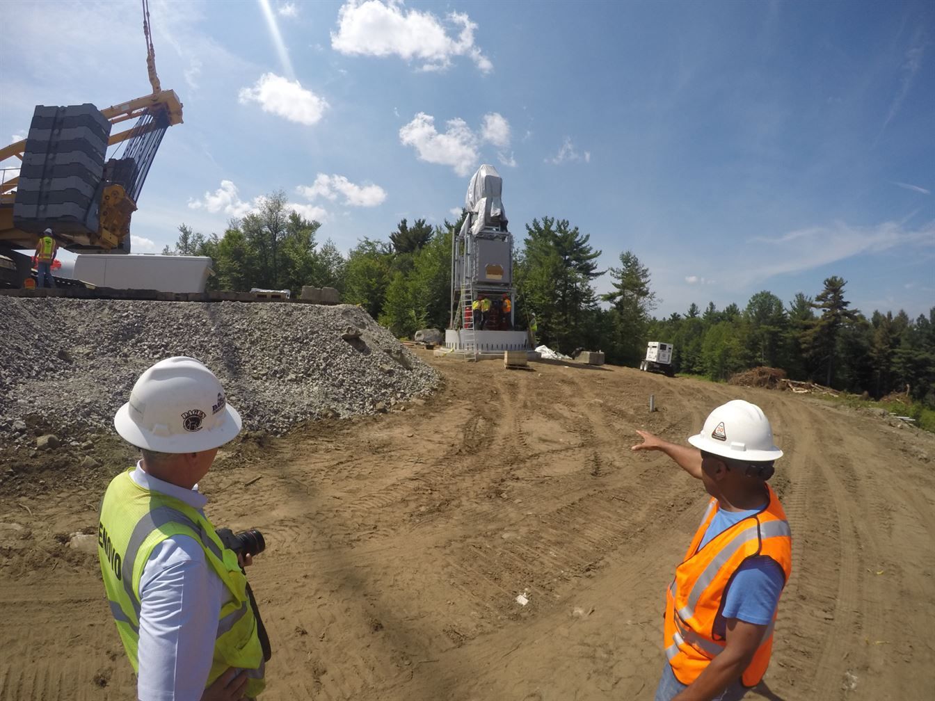 Two construction workers are standing in front of a large wind turbine.