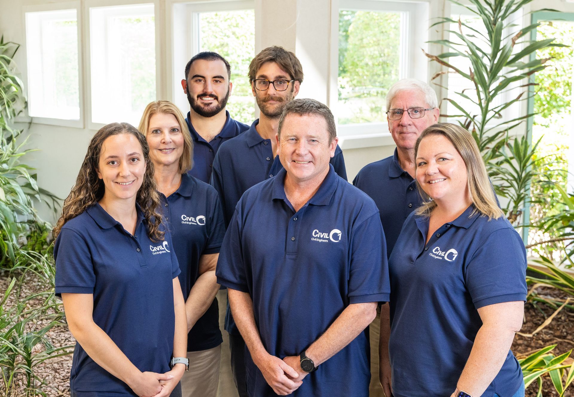 Group of people in matching blue shirts pose in front of a window, smiling.
