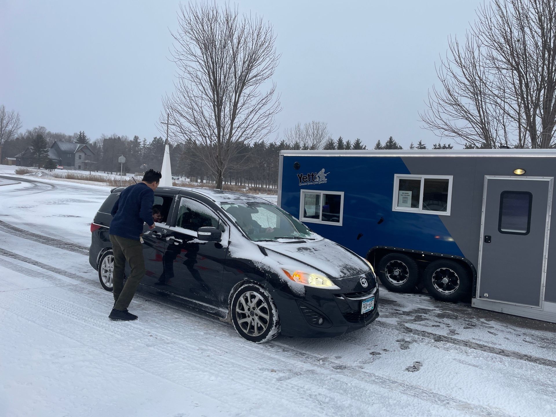 Black car towing a blue and gray trailer on a snow-covered road; a person stands by the car.