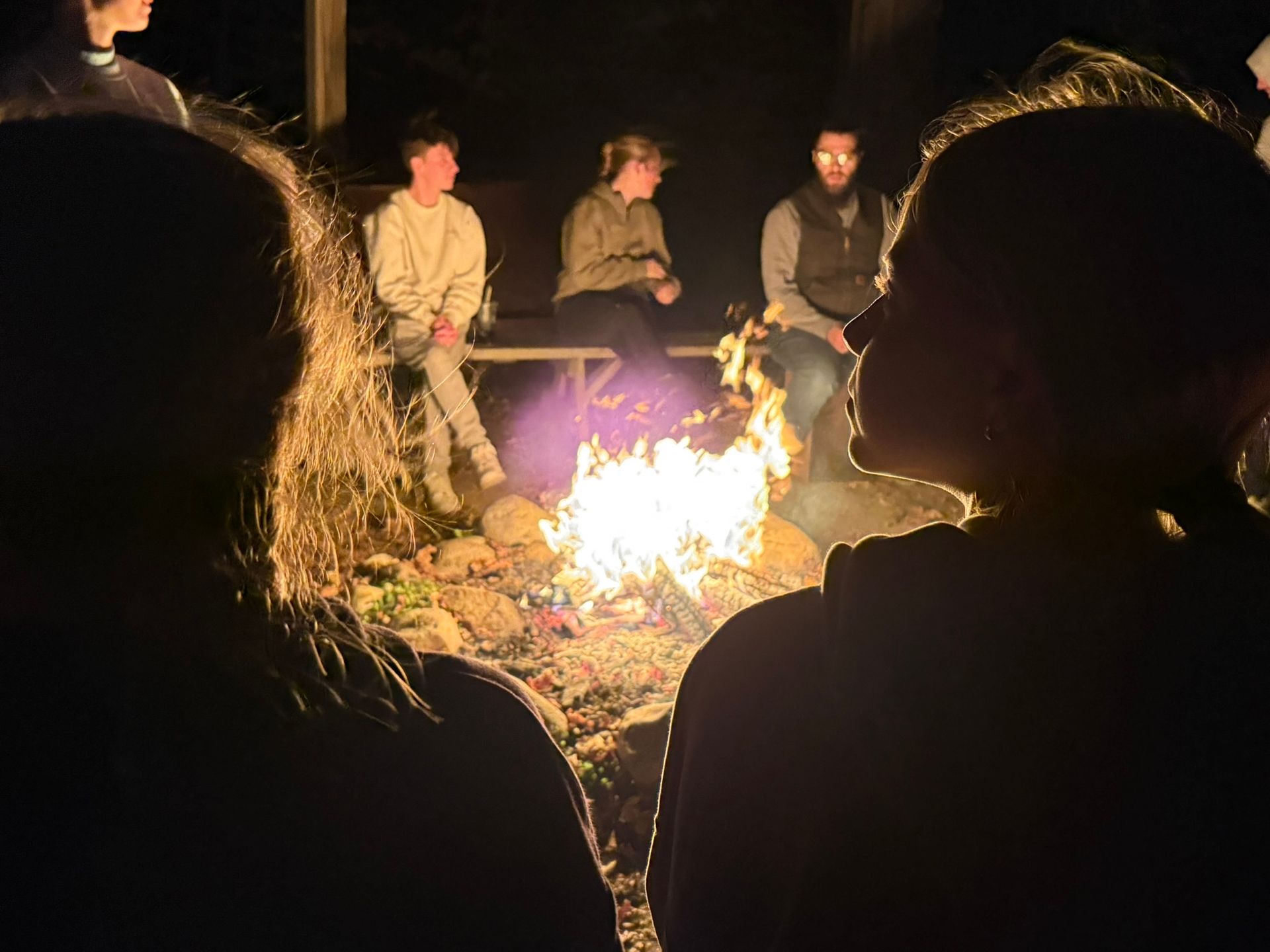 People gathered around a bonfire at night. Flames illuminate faces; others sit on a bench in the background.
