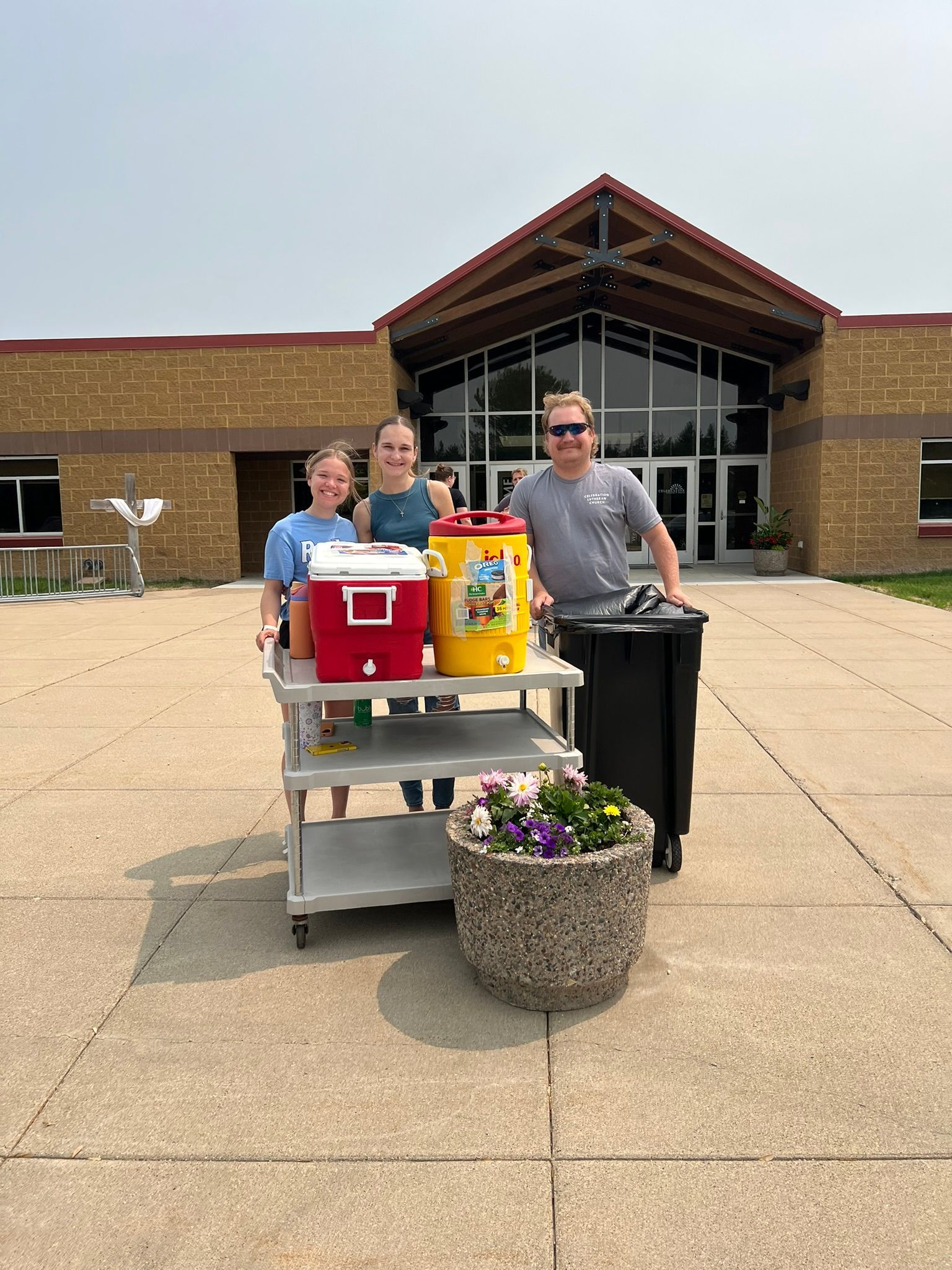 Three people behind a cart with red and yellow drink coolers outside a building entrance