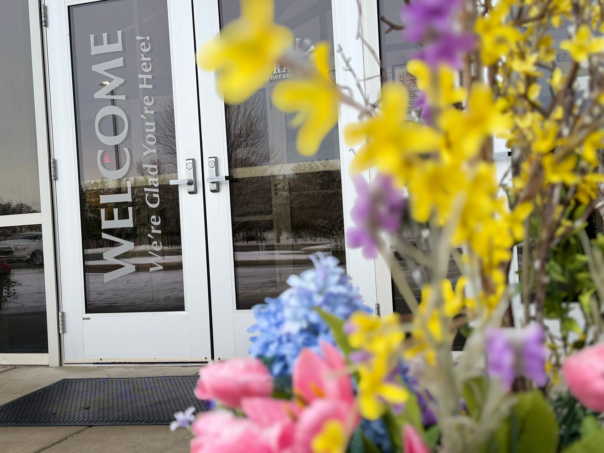 Lit building entrance with glass doors, illuminated at night. Two potted plants flank the entrance.