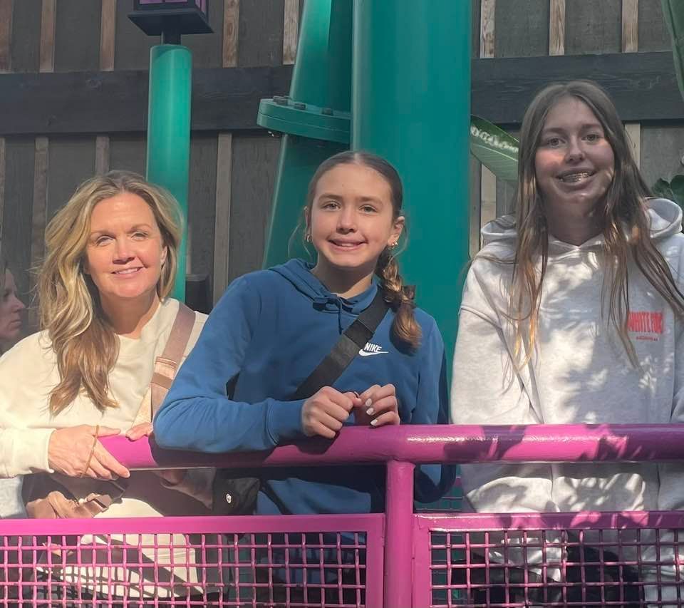 Woman and two girls smile, posing near a purple railing. Green machinery and structures are visible behind them.