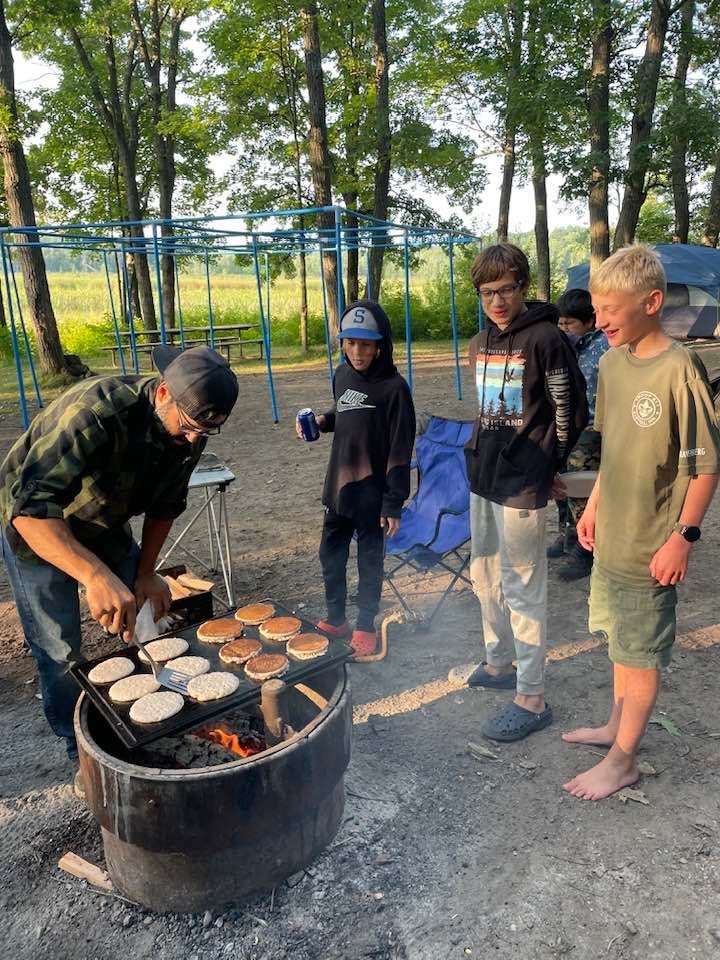 People gathered around a fire pit grilling burgers outdoors near a trampoline.