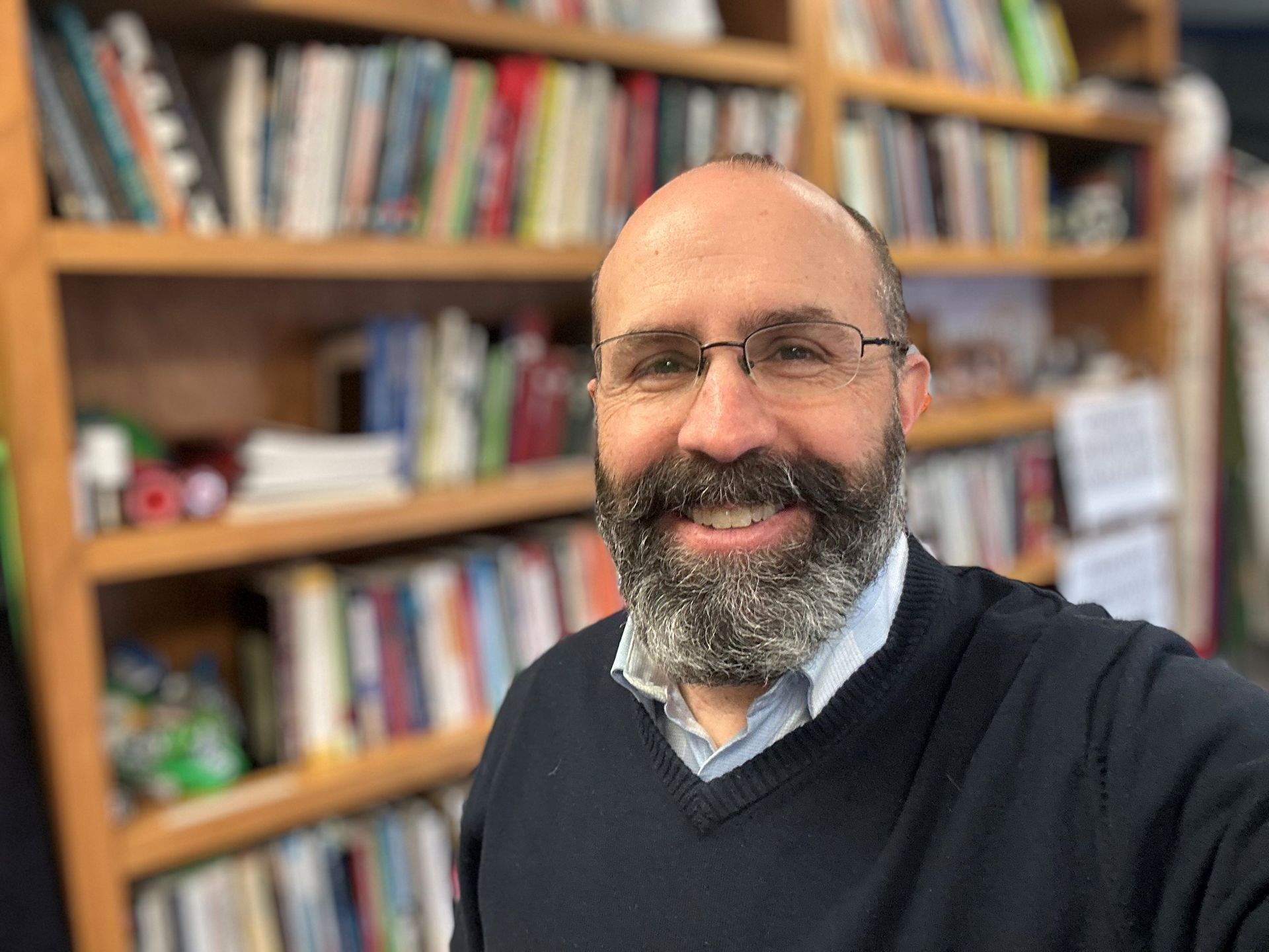 A man with a beard and glasses is smiling in front of a bookshelf.