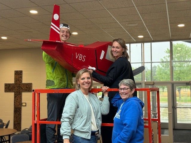 A group of people are standing around a model airplane that says vbs