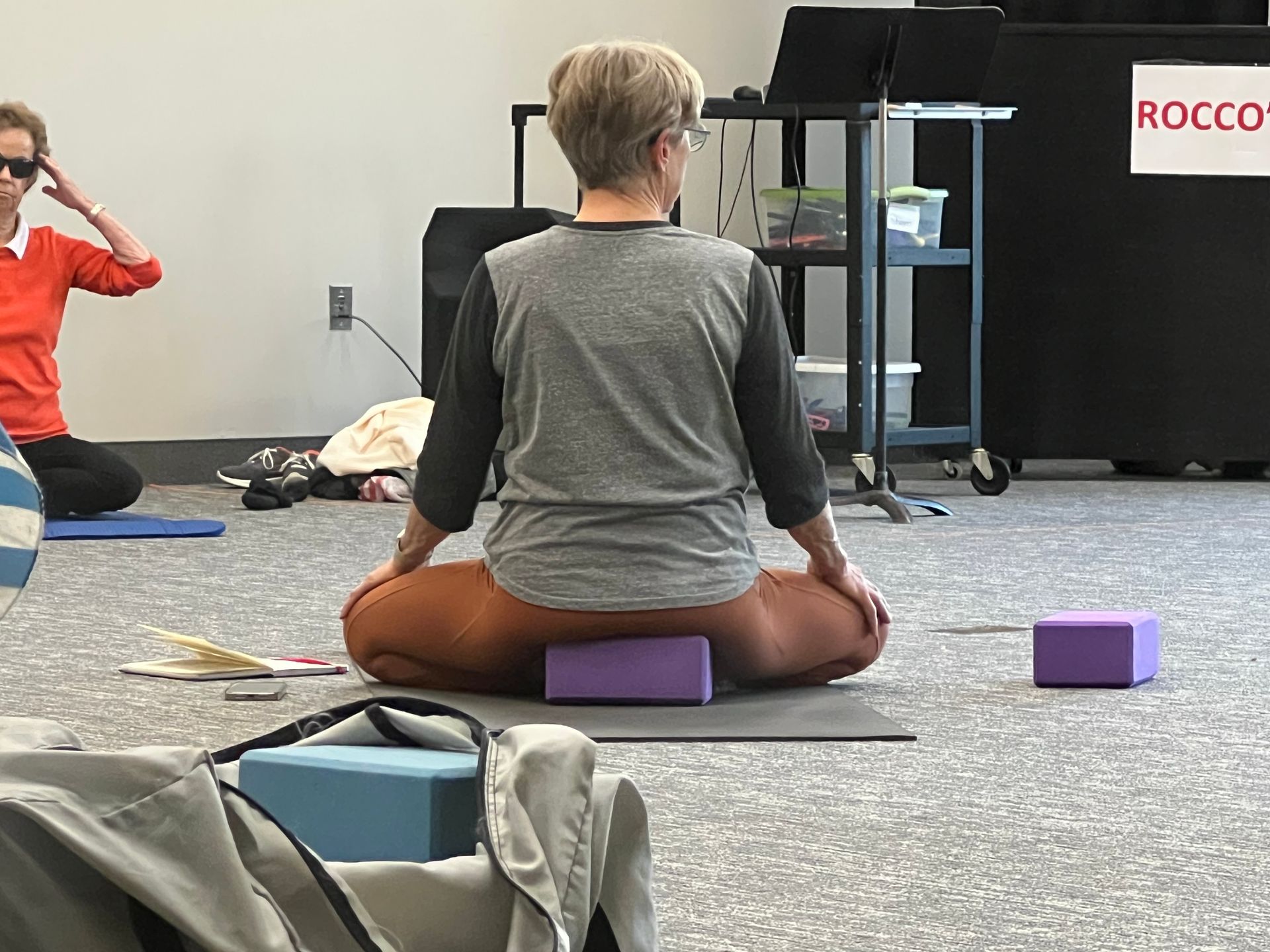 Person seated on a purple yoga block in a studio, facing a wall with other props nearby.