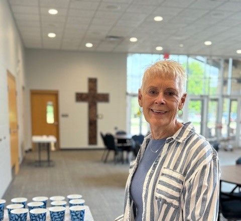 A woman in a striped shirt is standing in a room with a cross in the background.