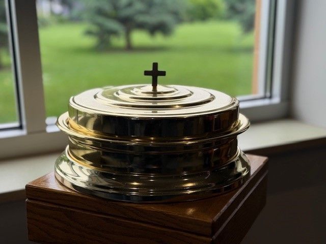 A brass container with a cross on top is sitting on a wooden box in front of a window.