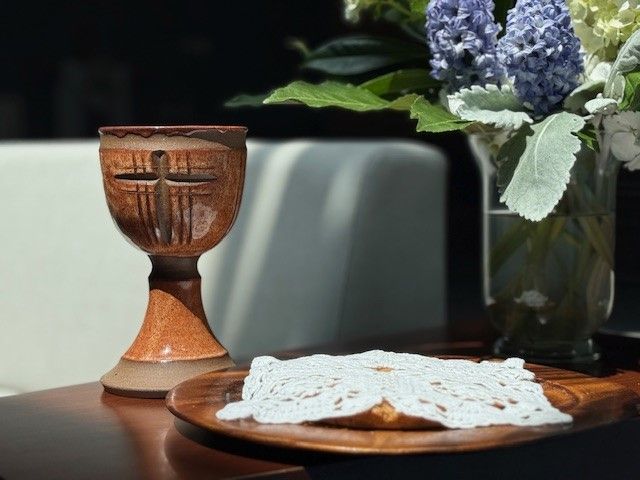 A wooden chalice with a cross on it sits on a table next to a plate of bread and a vase of flowers