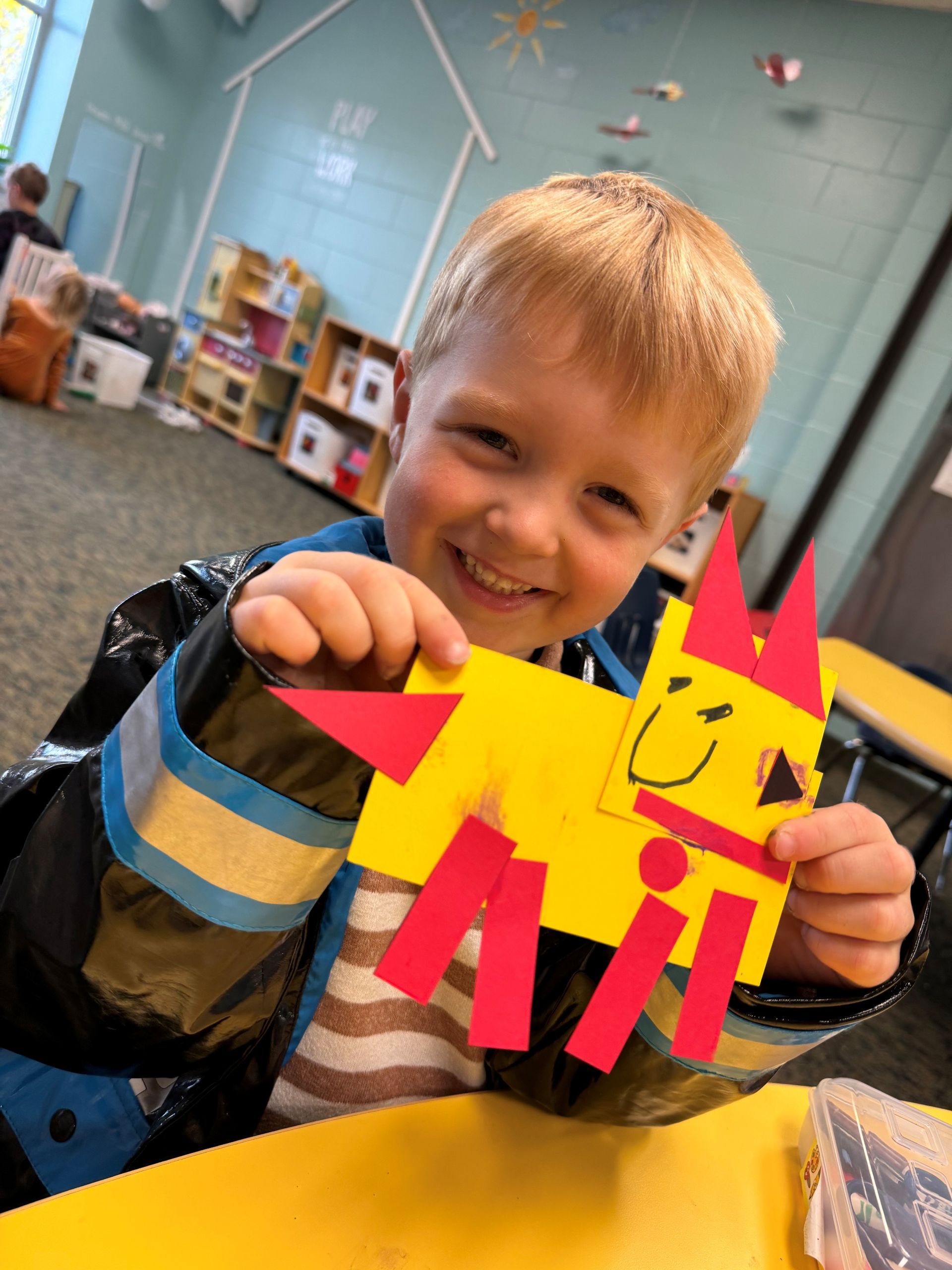 A young boy is sitting at a table holding a yellow and red craft.