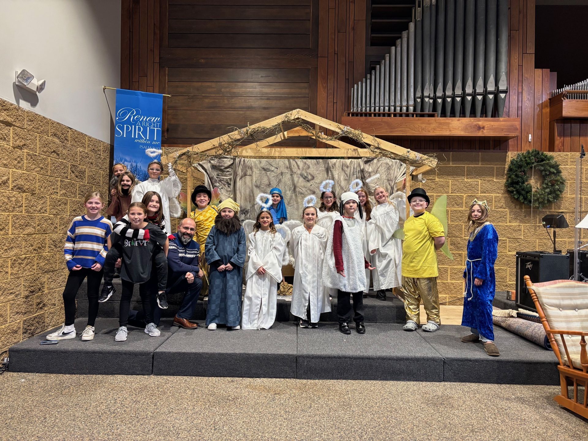 Children in costumes pose on stage in front of a nativity scene backdrop in a church.