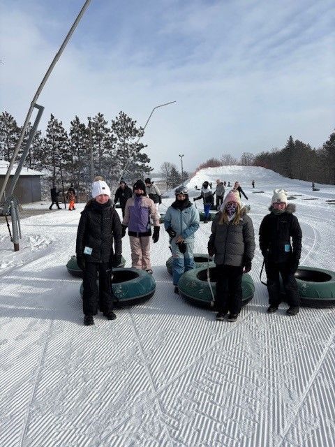 People in winter clothing stand with inner tubes on a snowy hill, ready for tubing.