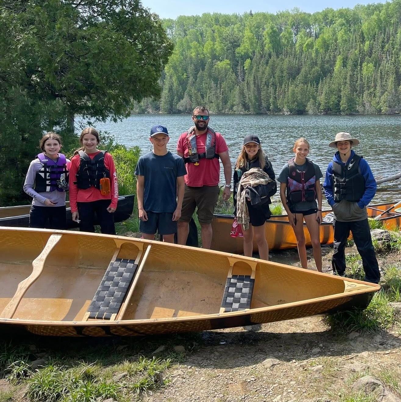 A group of people are standing next to a canoe on the shore of a lake.