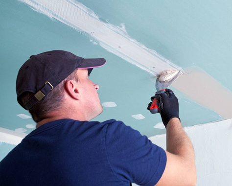 Man in cap applies joint compound to drywall ceiling seam with taping knife.