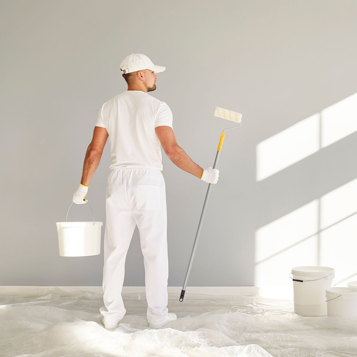 Man in white paint suit and cap holding paint roller and bucket, painting a gray wall.