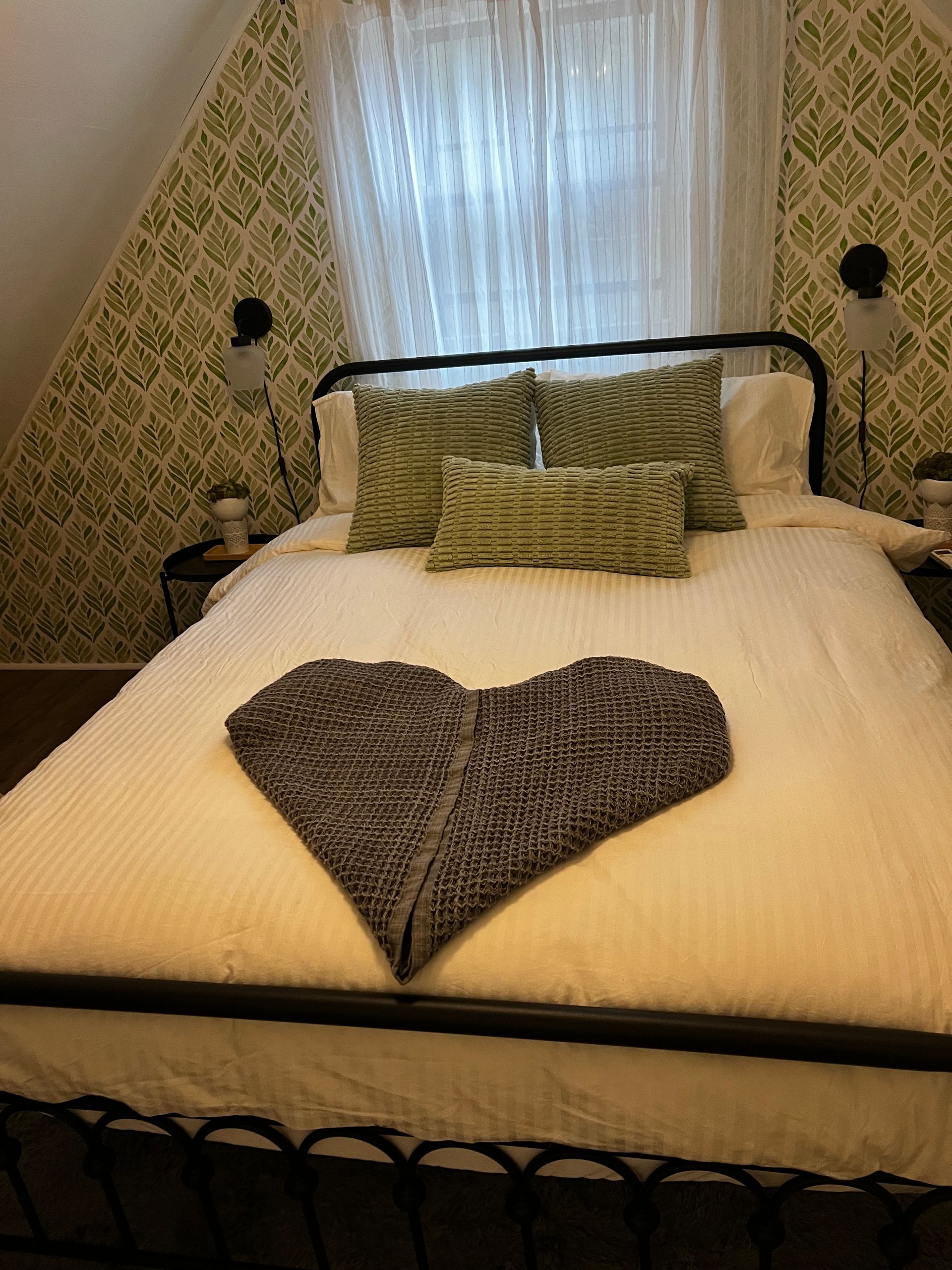 Cozy bedroom with a bed made with white linens and green pillows, and a gray blanket folded in a heart shape.