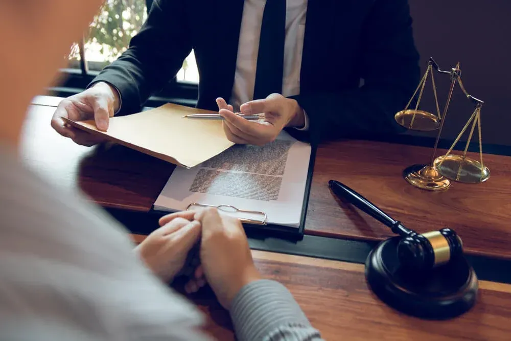 Lawyer handing documents to a client at a wooden table. Gavel and scales of justice are present.