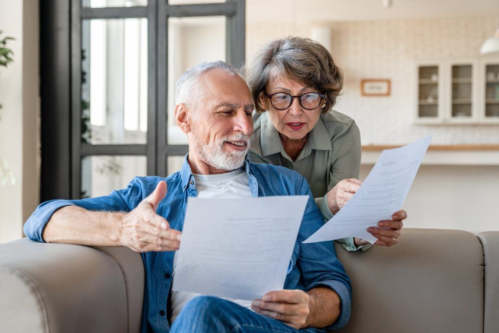 Two people sit on a couch in a brightly lit room, looking at documents they are holding and discussing them intently.