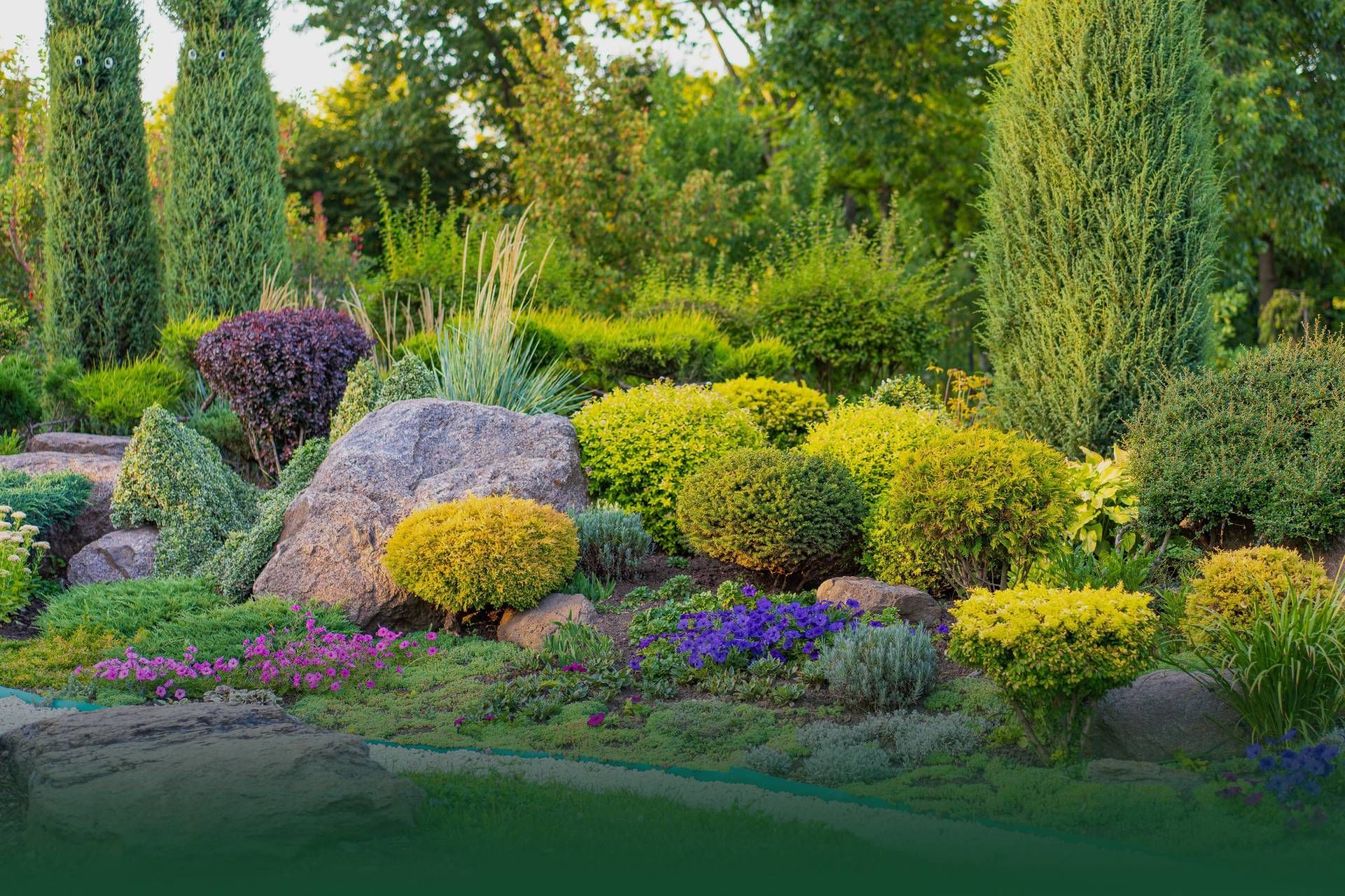 Lush, landscaped garden with various shrubs, flowers, and tall, green trees. Rocks are prominent.