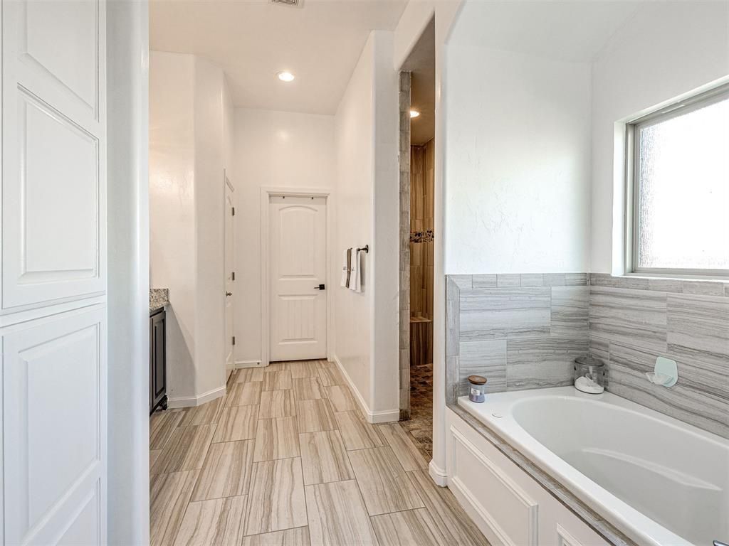Bright bathroom with a long hallway, white walls, wood floor, and a bathtub with tiled surround.