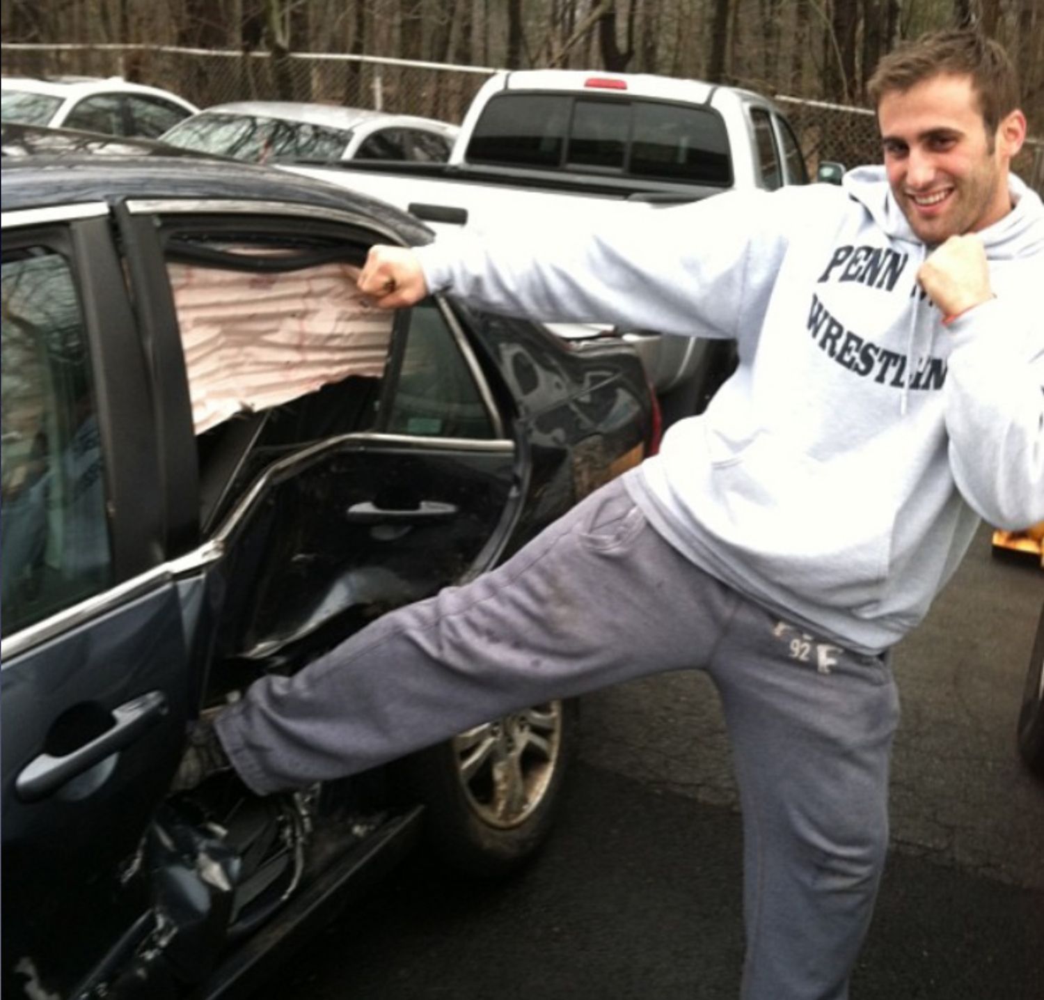 A man wearing a penn wrestler sweatshirt is kicking a car