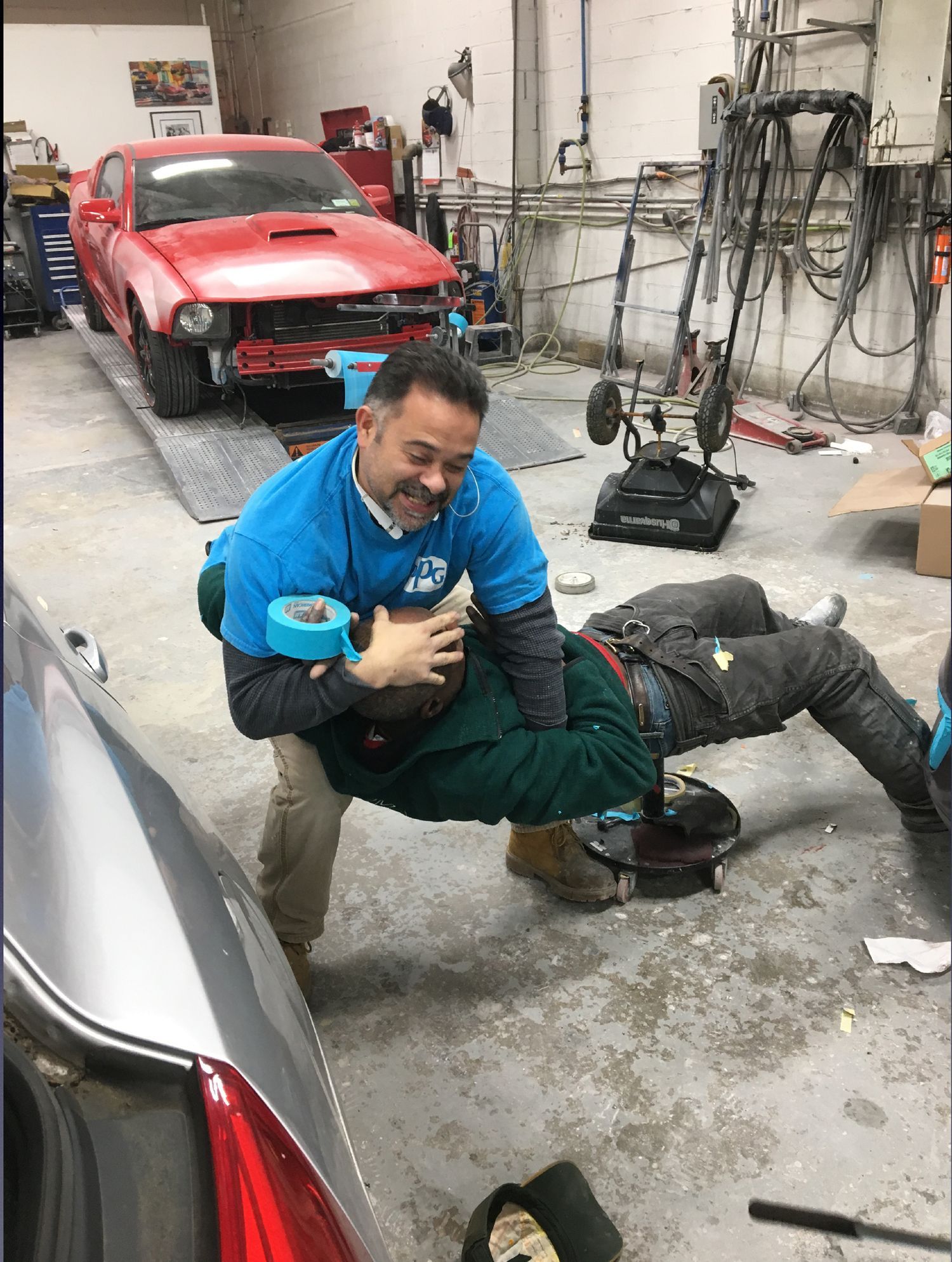 A man in a blue shirt is helping another man in a garage.