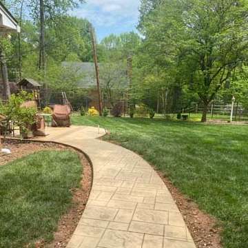 A stamped concrete walkway curves through a green lawn, leading toward a house in a wooded setting