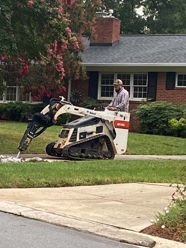 Man operating a Bobcat with a jackhammer attachment, breaking up concrete in front of a brick house