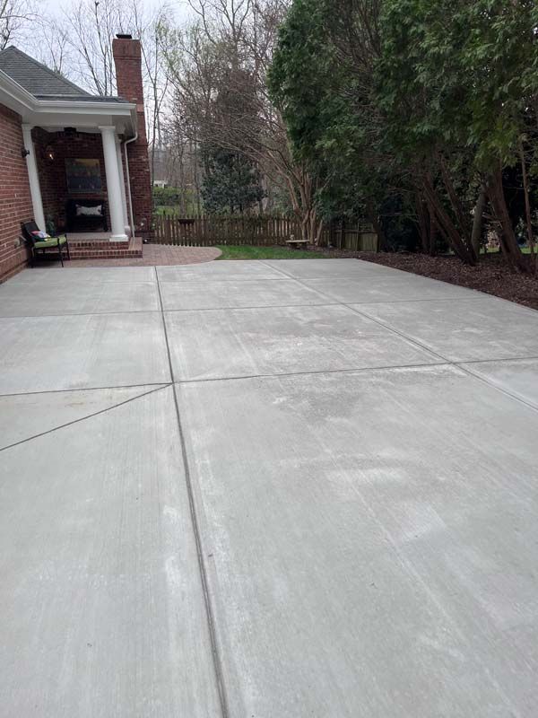 Large concrete patio next to a brick home with a covered porch, trees, and grass