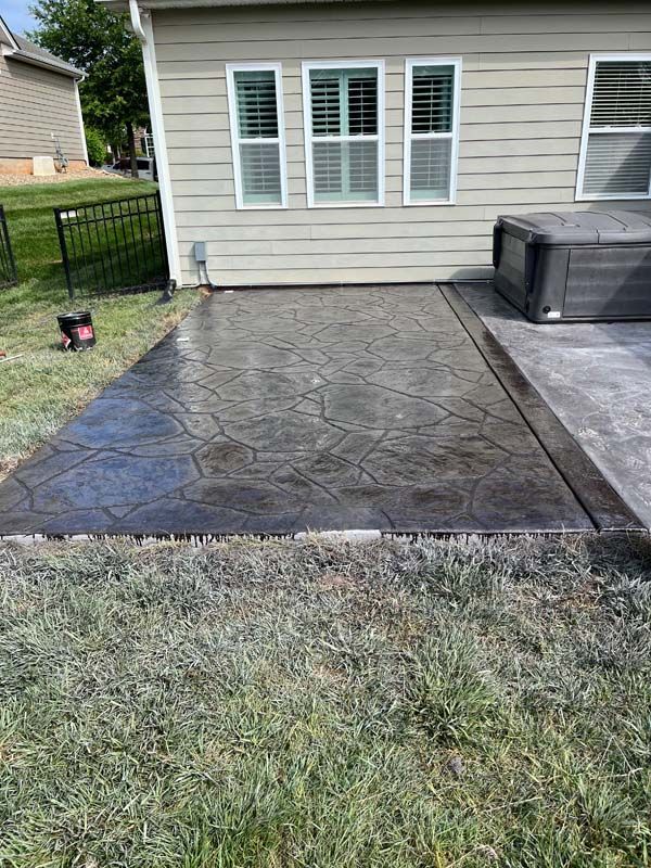 A dark, patterned concrete patio next to a house with three windows and a hot tub