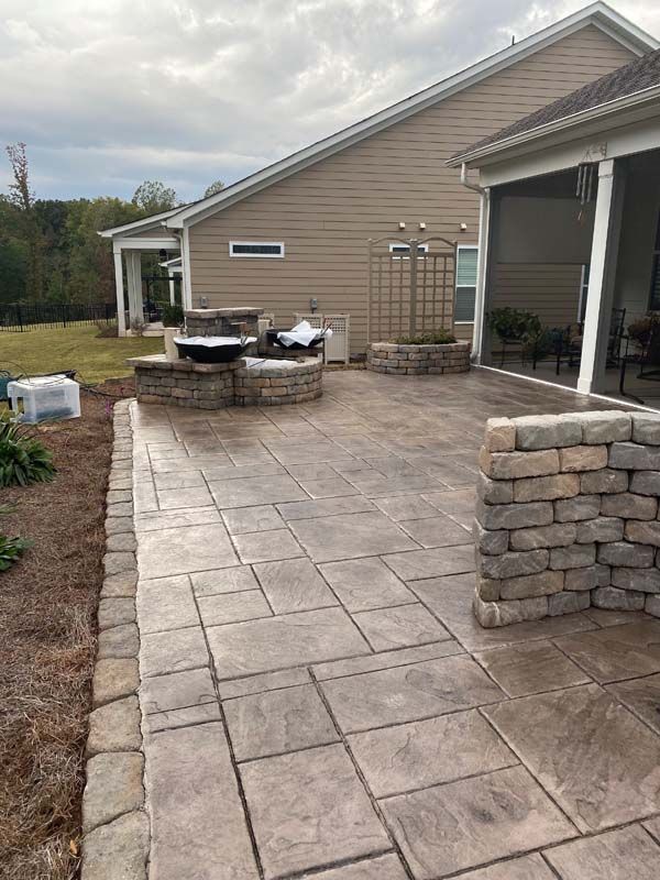 Stone patio with decorative wall, water feature, and fire pit beside a tan house under a cloudy sky