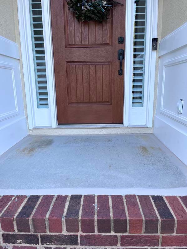 Brown front door with wreath, concrete porch, brick step, flanked by white trim with shutters