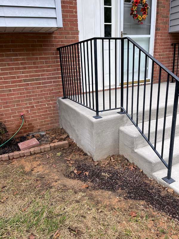 Brick house entrance with concrete steps, black railing, and a small flower bed