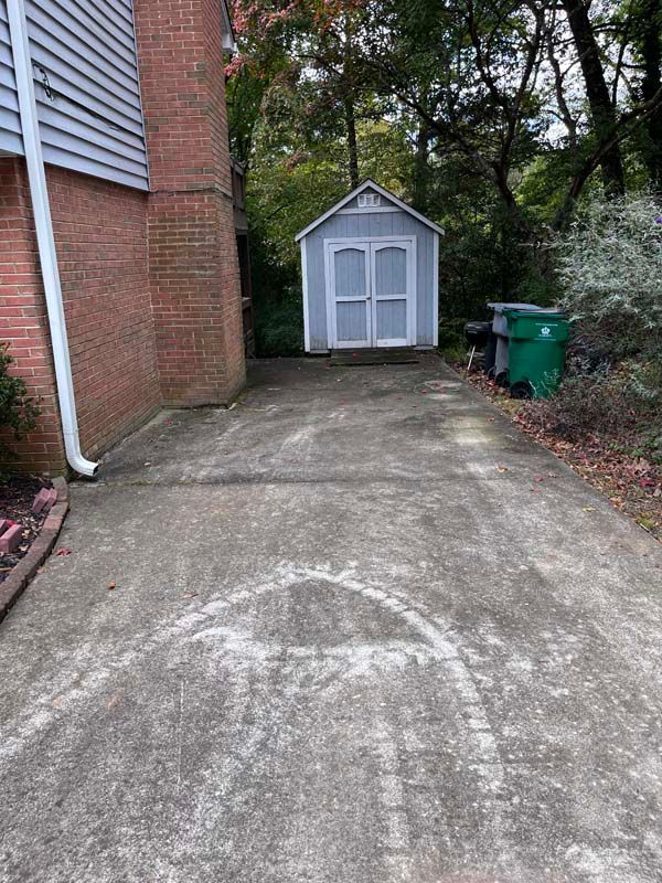 Concrete driveway leading to a light blue shed. Brick building on the left, green trash bin on the right.