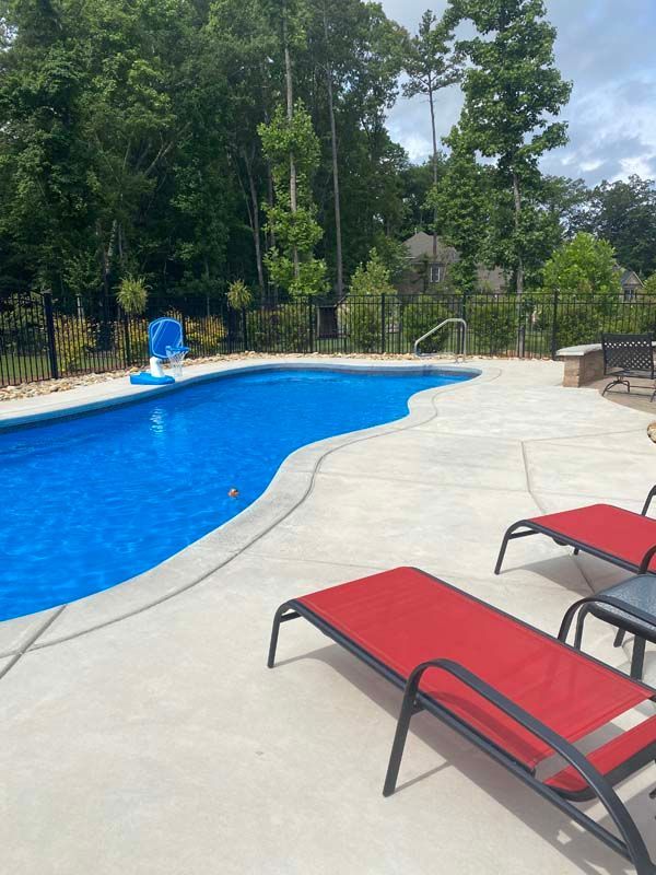 A backyard pool with blue water and lounge chairs with red cushions on concrete patio