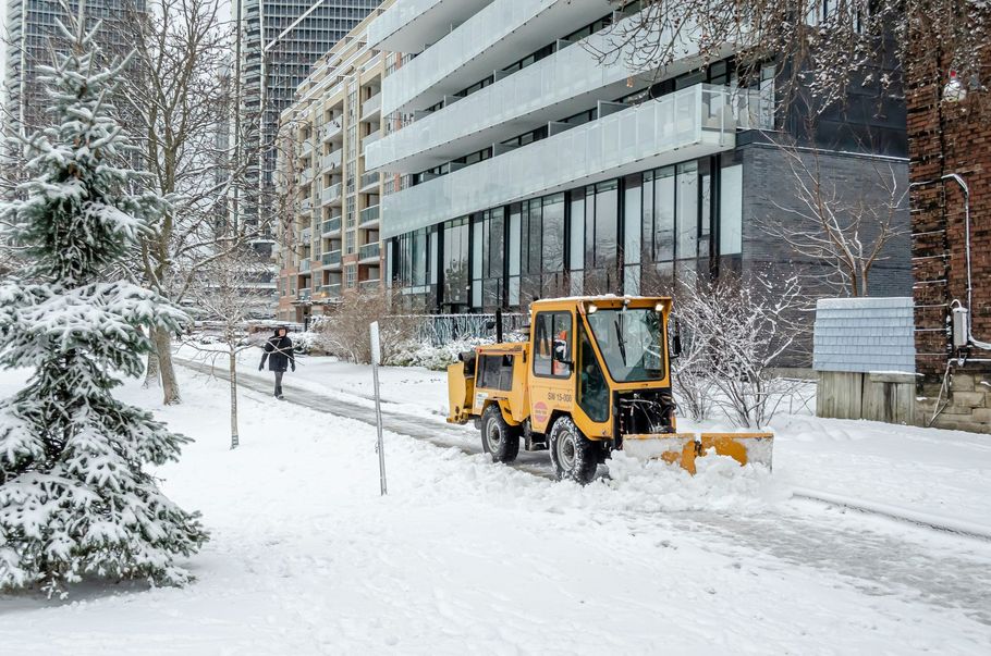Yellow snowplow clearing a snowy sidewalk beside a modern building
