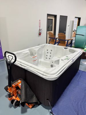 A new, square hot tub, with dark brown siding and a white interior, sits next to a pallet jack indoors.