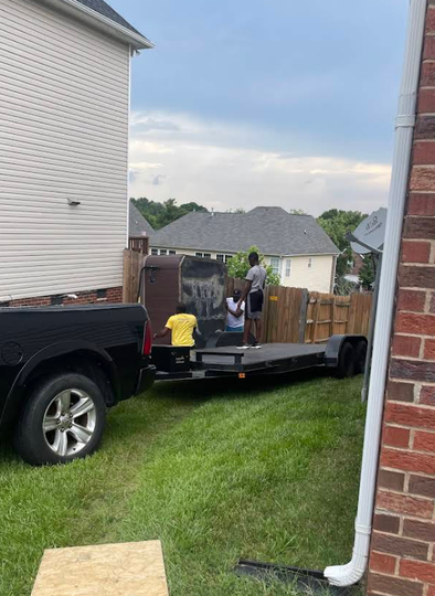 People loading an item onto a trailer, hitched to a black pickup truck, in a grassy yard between houses.