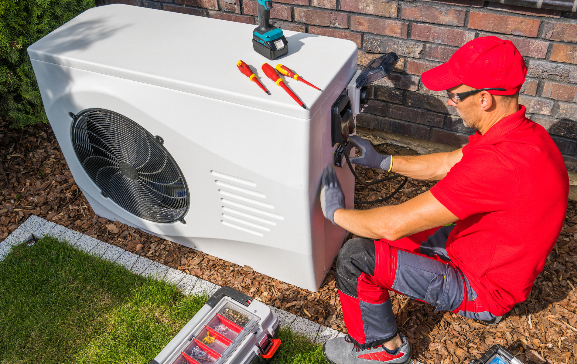 A man is sitting on the ground working on a heat pump.